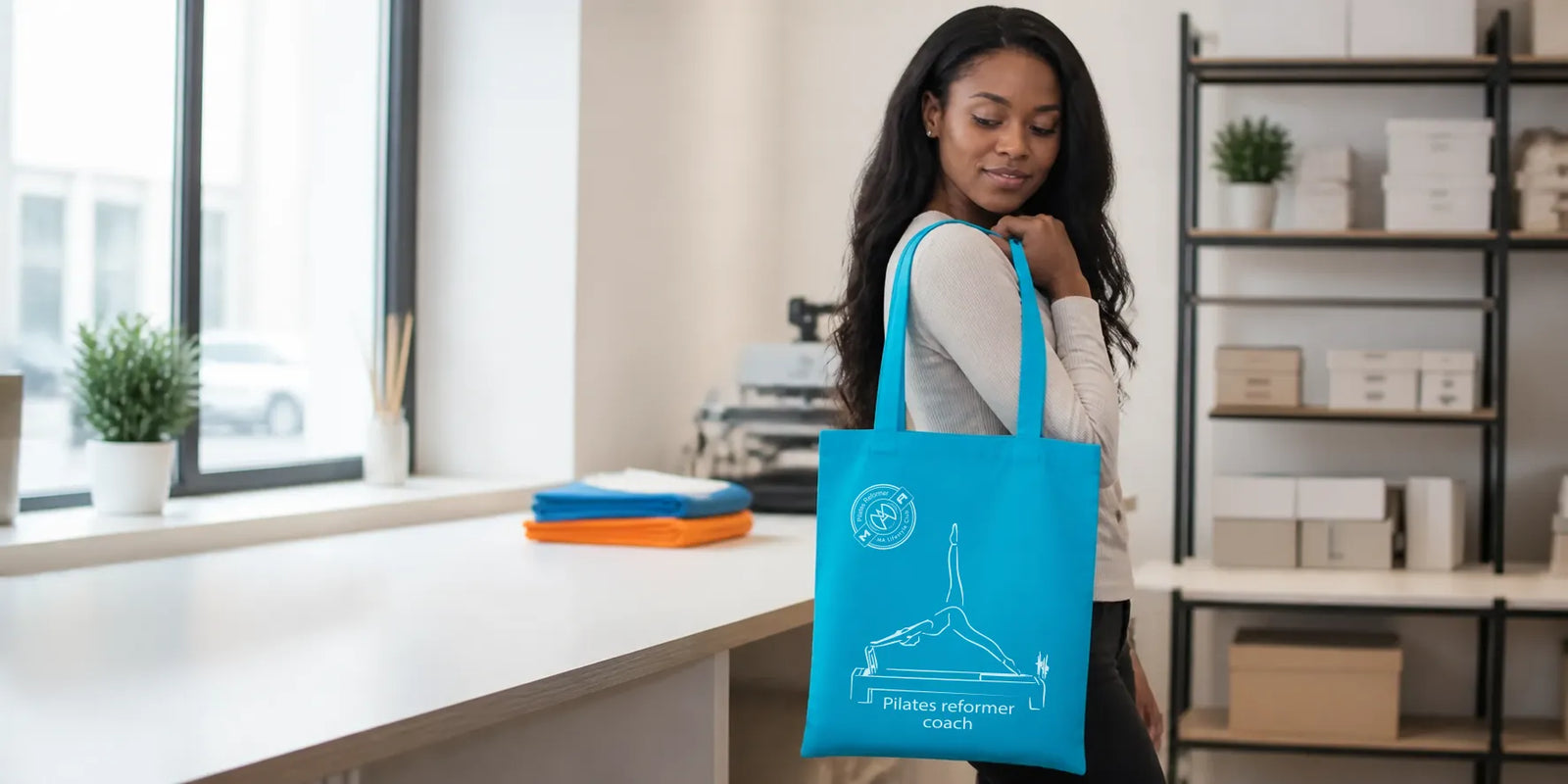 Vrouw met turquoise draagtas met wit logo in een lichte werkruimte.